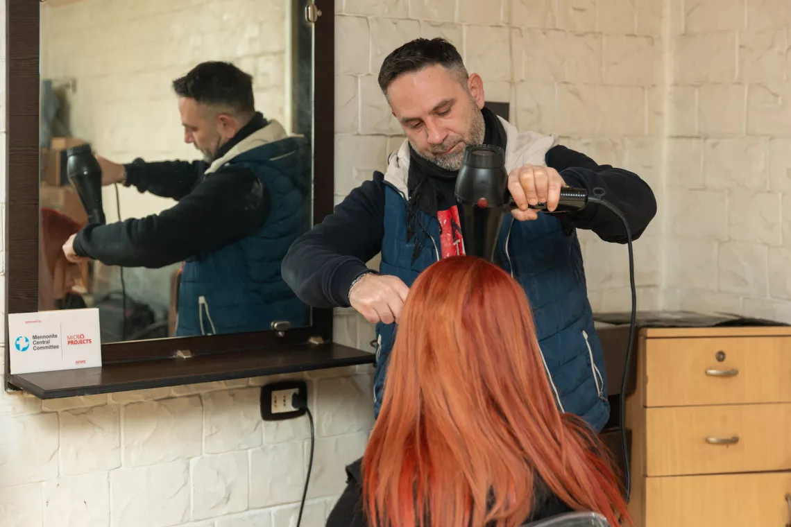 A hairdresser is styling a client's red hair with a blow dryer, reflected in a mirror in a salon setting.