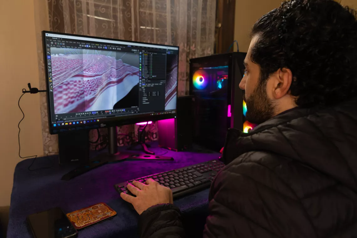 A person is working on a 3D modeling software on a computer with dual monitors. The room is dimly lit, and the computer has RGB lighting.