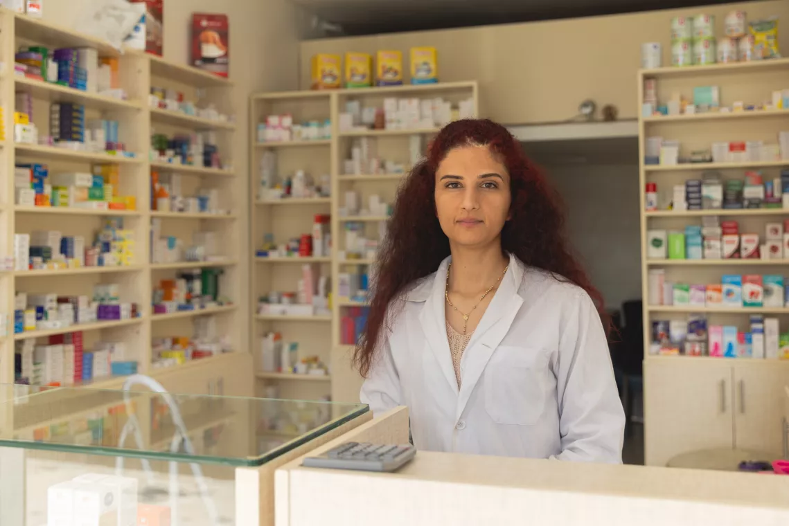 A woman in a white coat stands behind a pharmacy counter with shelves of medications in the background. She appears professional and attentive.