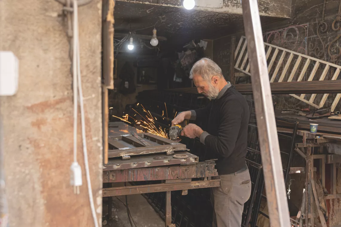 A man is working with metal in a workshop, surrounded by tools and materials. Sparks fly as he focuses on his task, suggesting metal grinding or welding.