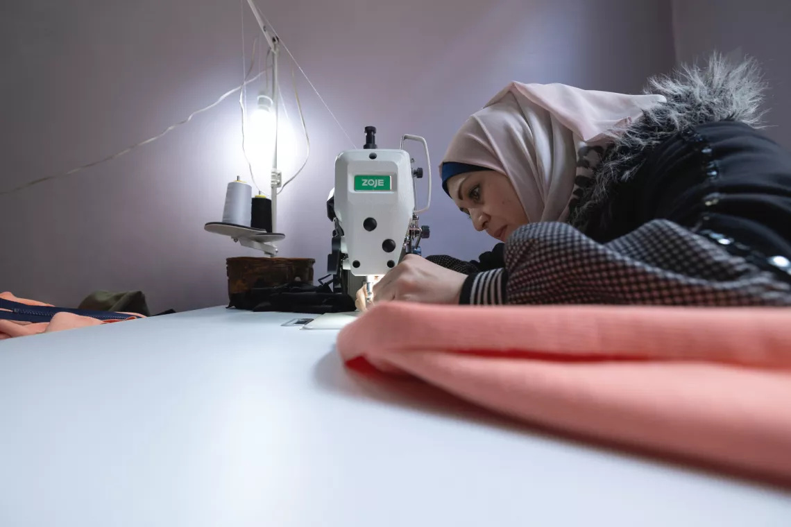 A woman wearing a hijab is focused on sewing with a machine, working on a piece of red fabric in a dimly lit room.