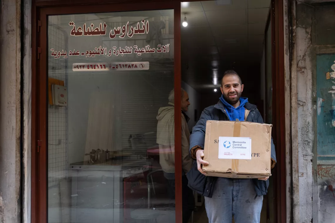 A man stands in a doorway holding a cardboard box with a logo on it. He smiles at the camera. Arabic text is visible on the door's glass pane.