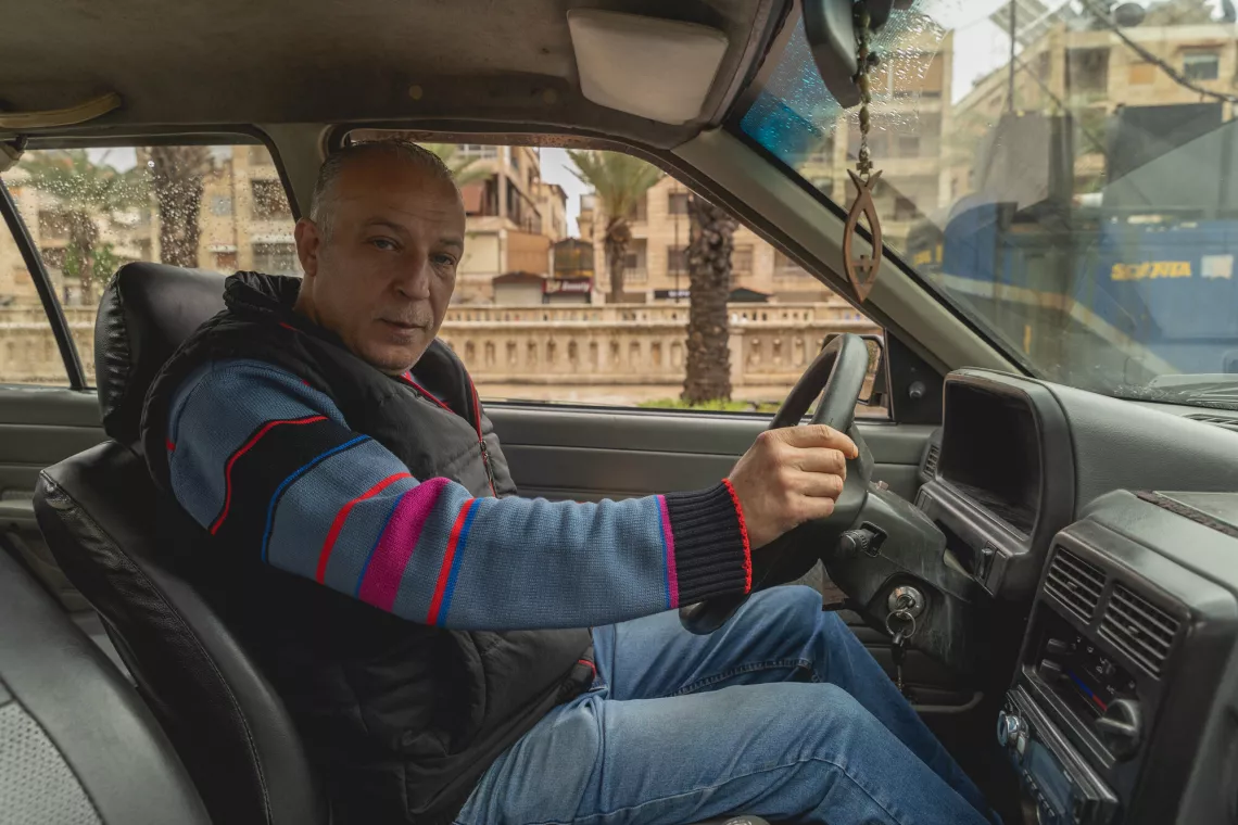 A man is driving a car, looking at the camera. He wears a striped sweater and jeans. The car's interior and a blurred street scene are visible through the windows.