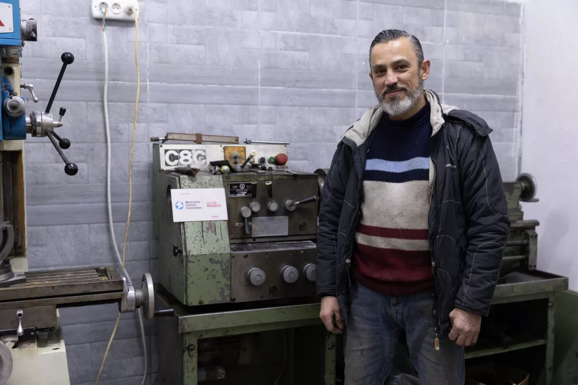 A man stands in a workshop with machinery, wearing a jacket and striped sweater, looking at the camera with a slight smile.