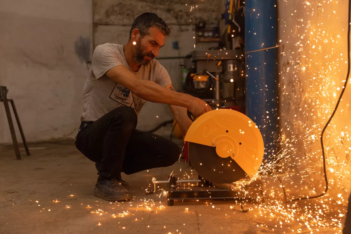 A man is crouching while using an angle grinder on metal, causing bright sparks to fly in a workshop setting.