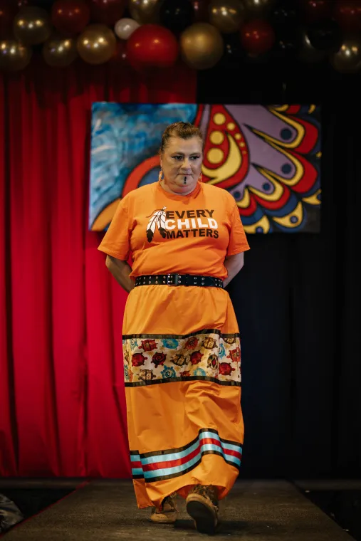 A woman wearing an Every Child Matters t-shirt models a multi-colored ribbon skirt.