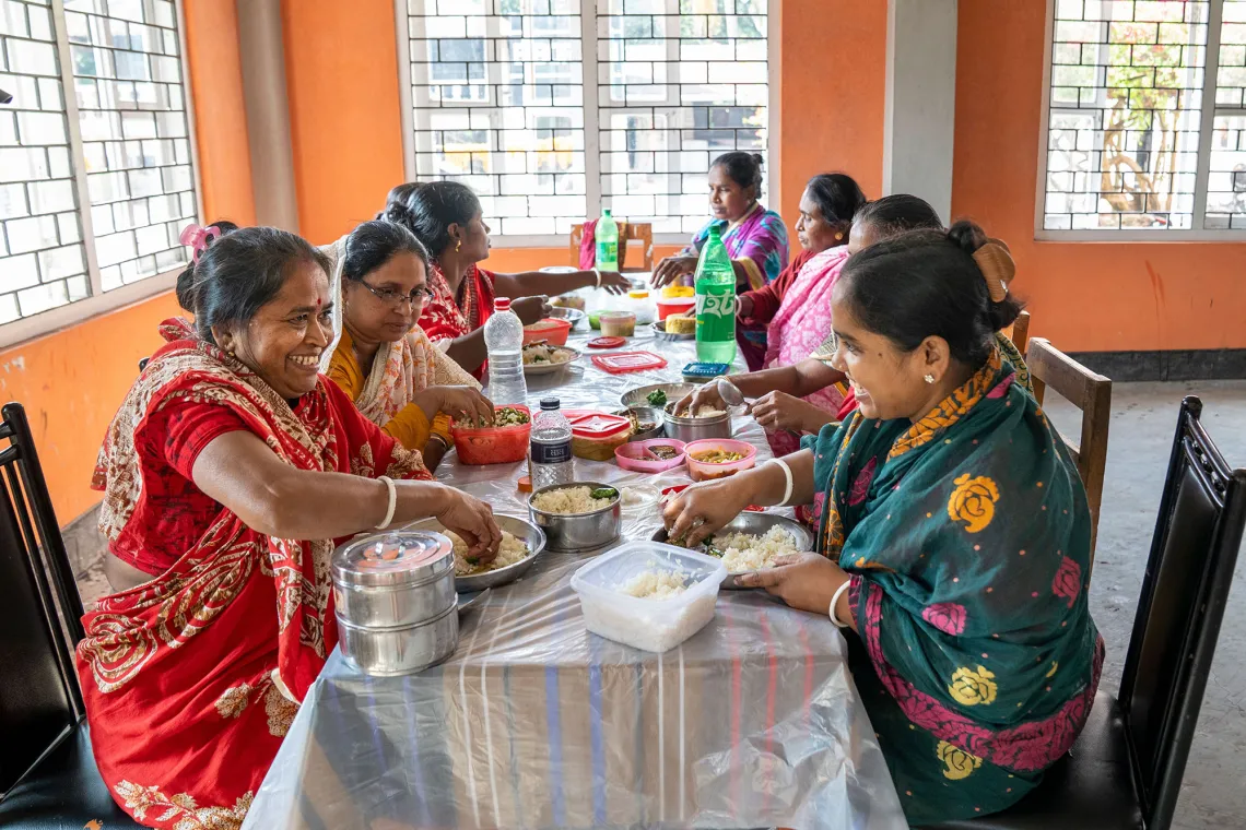 Seven women sit across from each other at a long table, having a meal.