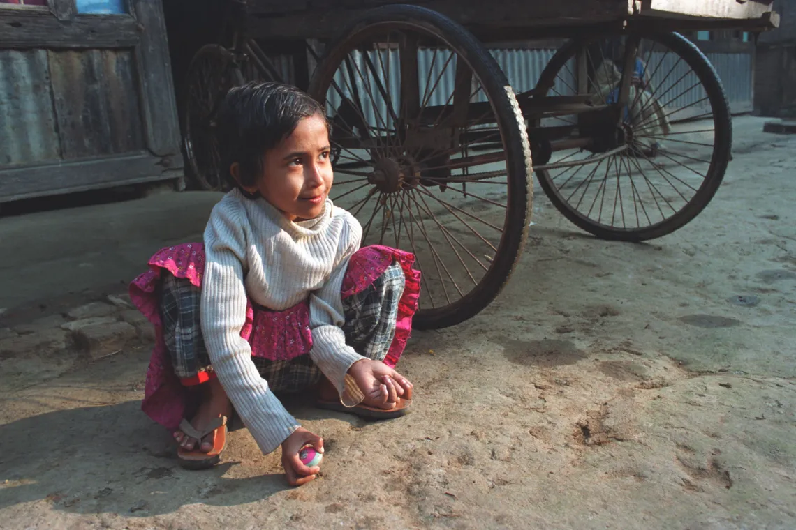 A young child squats on the ground outdoors with a toy in her hand.
