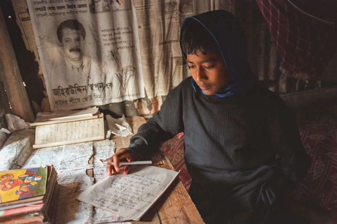 A kid sits at a desk lit by a lamp writing a paper.