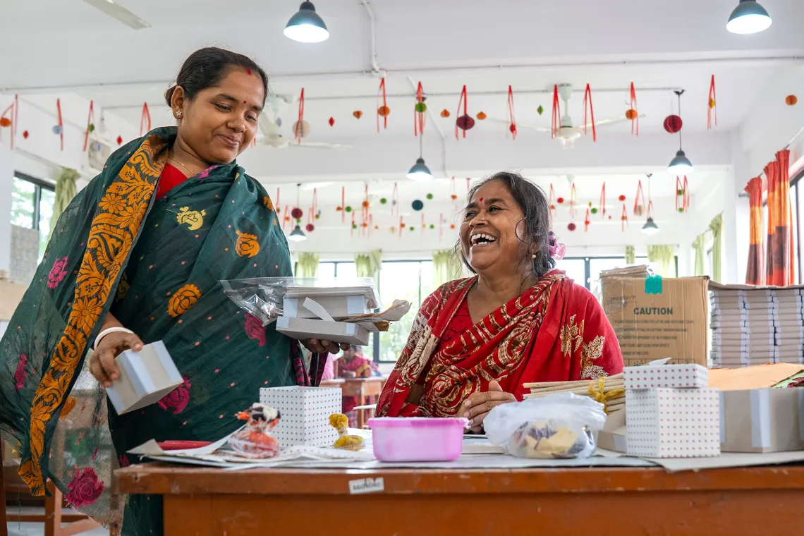 Two women, one standing and one sitting at a desk, enjoy a moment at a desk covered in paper products.