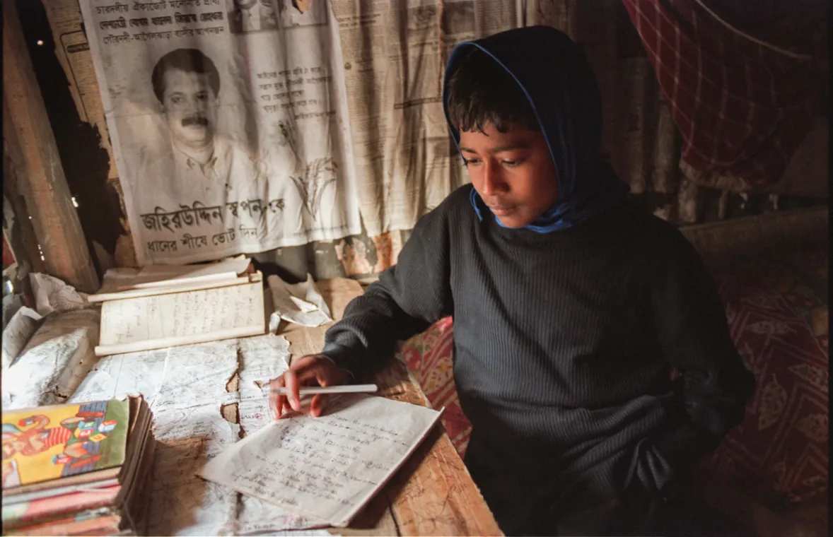 A child sits at a table, writing on paper surrounded by books and papers, with a poster on the wall in a modest indoor setting.
