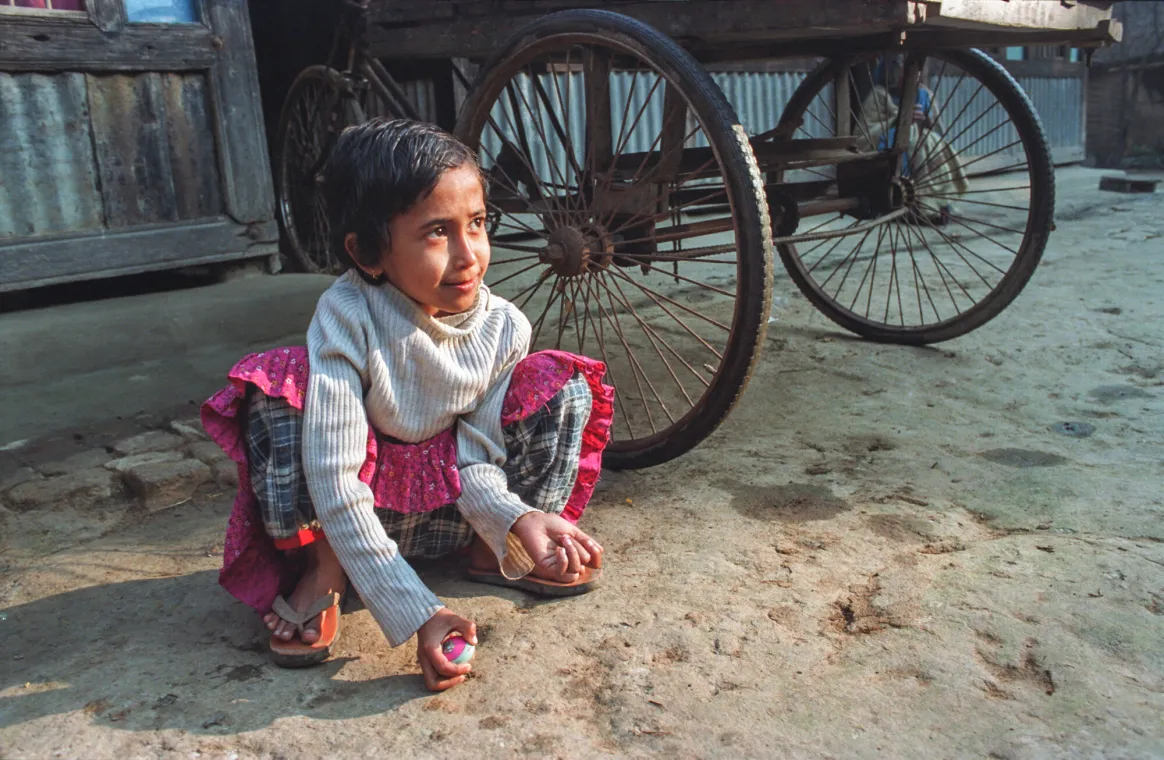 A young girl sits on the ground, holding an object, with a cart in the background and a textured surface beneath her.