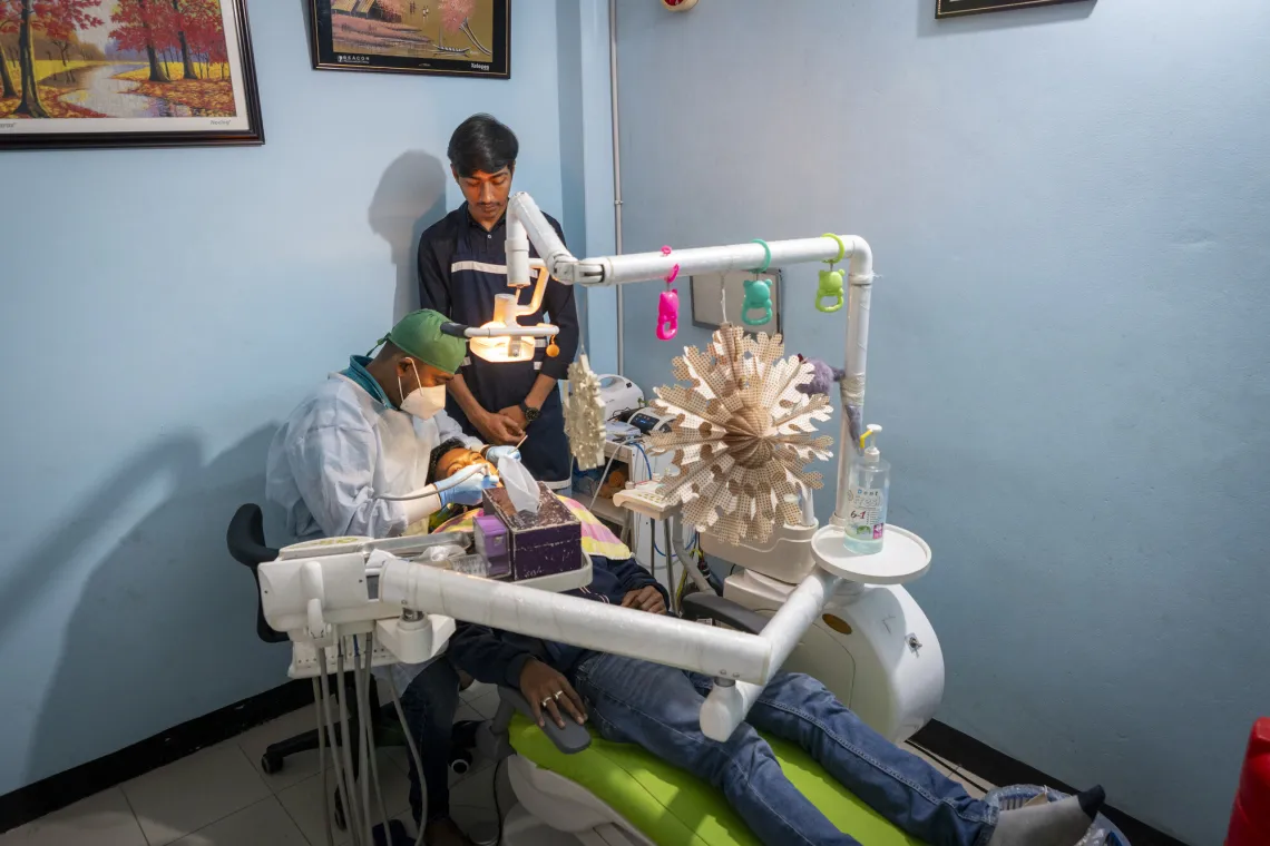 A dental office scene with a patient in a chair, a dentist working, and an assistant observing. Medical equipment is visible.