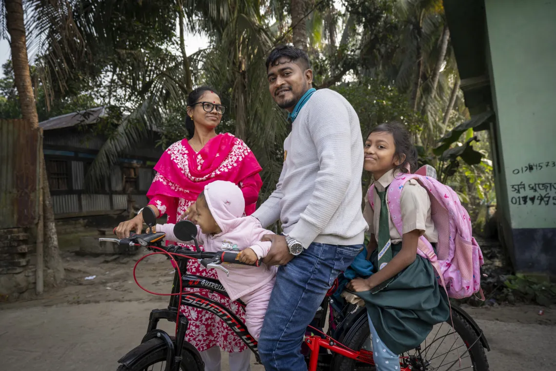 A man sits on a bicycle with two girls and a woman nearby. One girl carries a backpack, and a baby is seated in front. Trees are in the background.
