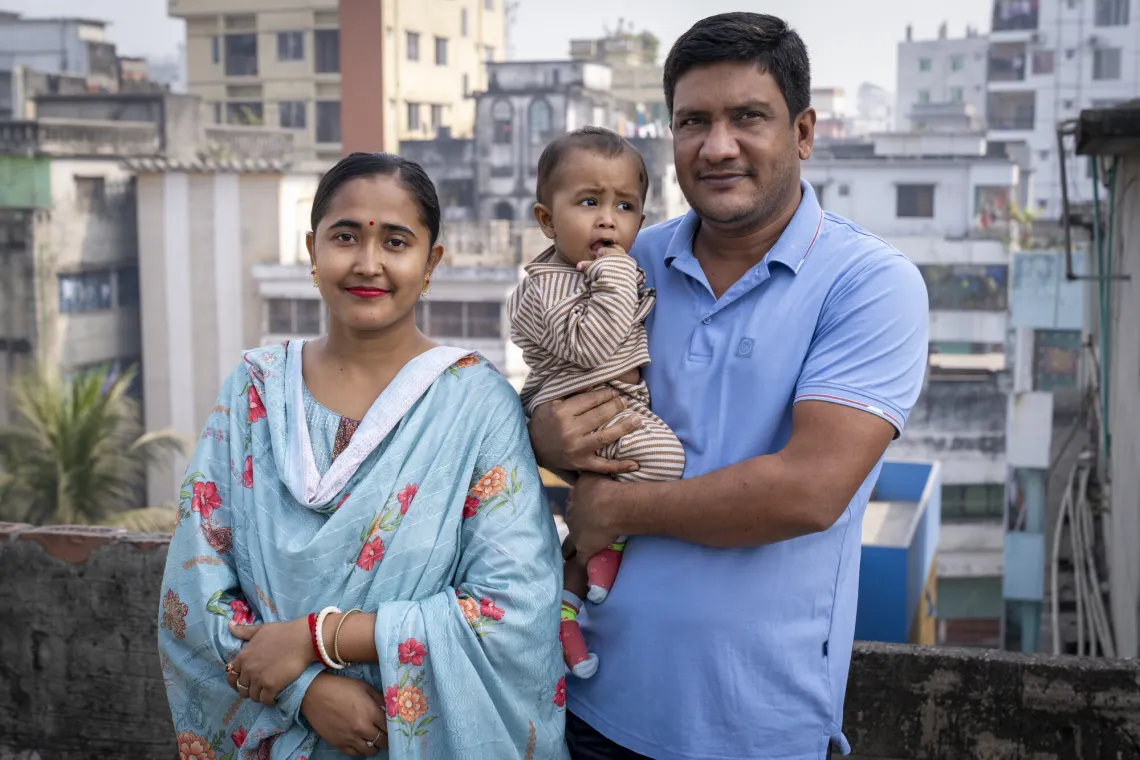 + A man holds a child while a woman stands beside them. They are outdoors with buildings and greenery in the background.