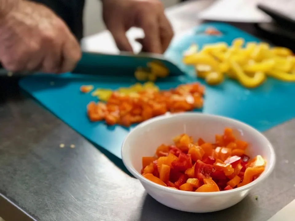 A pair of hands chopping orange bell peppers with red peppers in a bowl and yellow peppers on the other side of the chopping mat