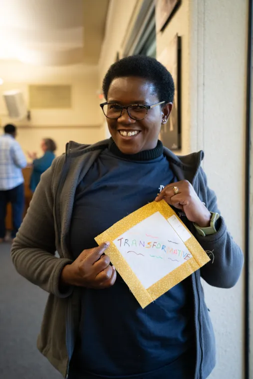 A person smiles while holding a decorated sign that reads "TRANSFORMATION." Others are visible in the background.