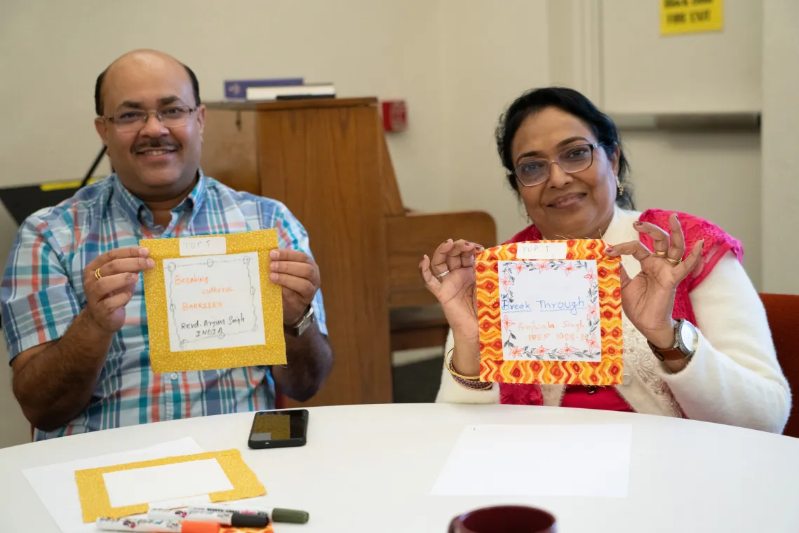 Two individuals sit at a table, each holding a decorated frame. A piano is visible in the background, along with art supplies.