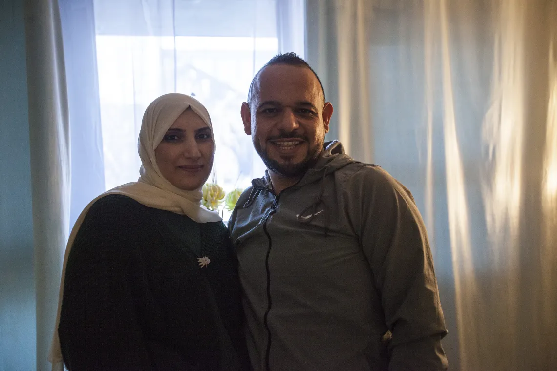 A Palestinian man and woman stand together and smile at the camera in an apartment