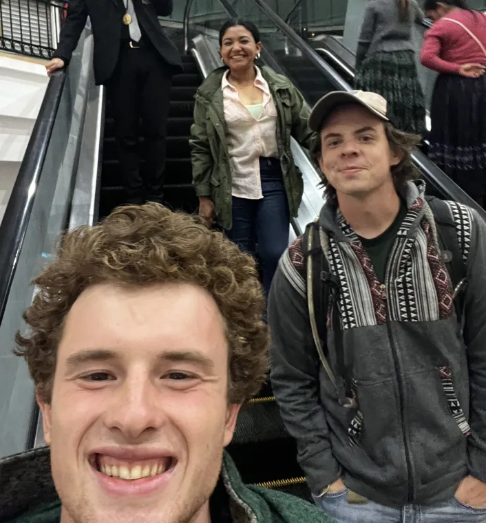 A group of three individuals on an escalator, with two men in the foreground smiling and one woman behind them.