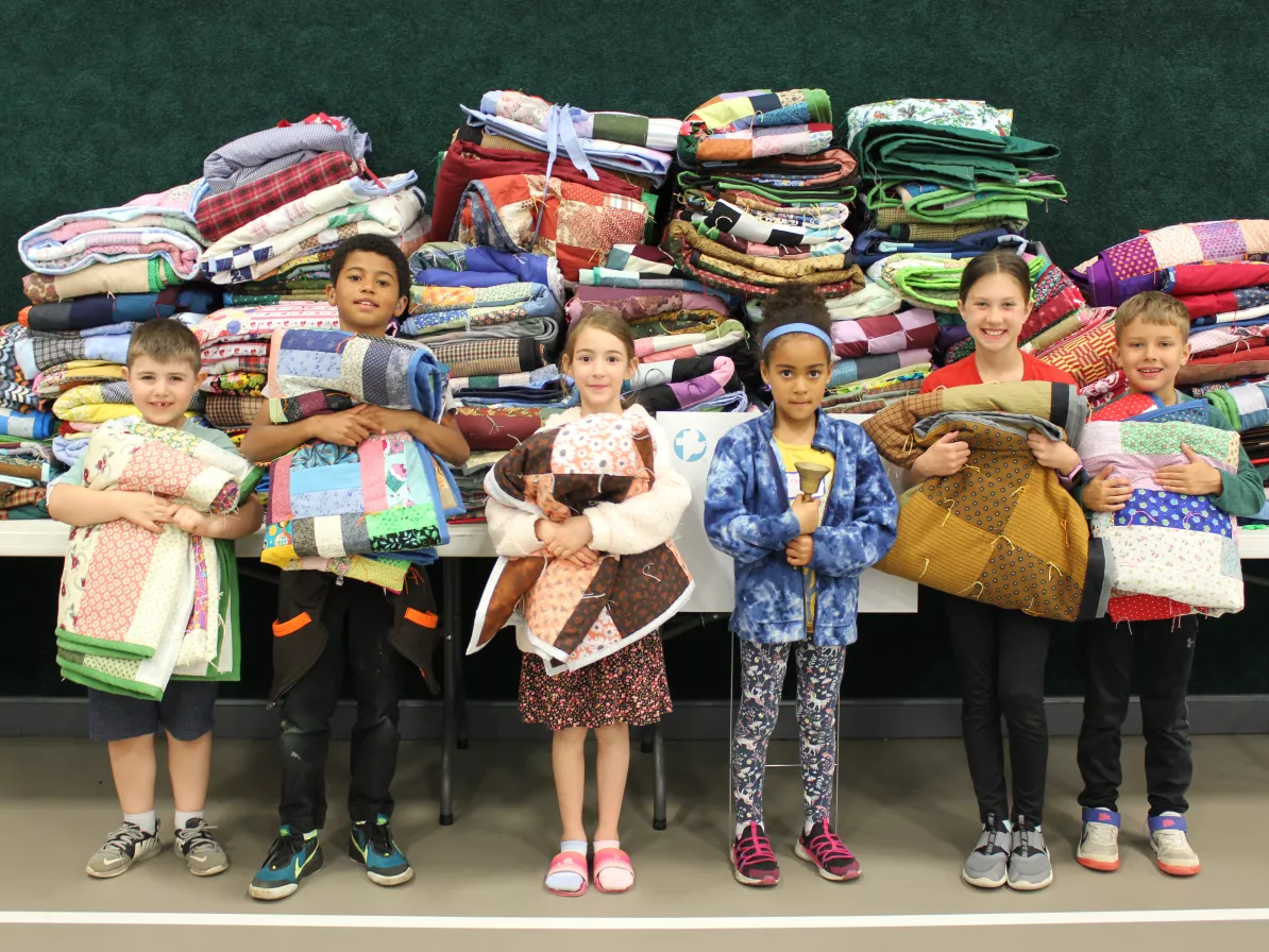 Six children stand in front a table laden with stacked comforters. They are all holding comforters too.