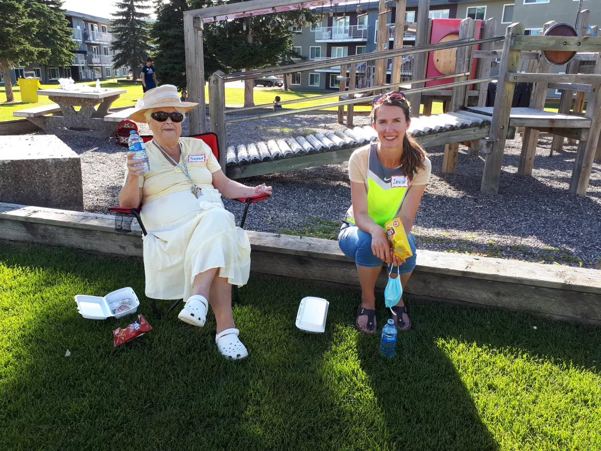 One woman sits on a lawn chair and holds a plastic bottle of water. Another woman sits next to her on the wooden ledge of a playground. She is holding a bag of chips.