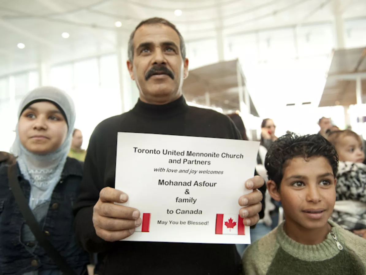 A family of three in an airport. The father holds a welcoming sign