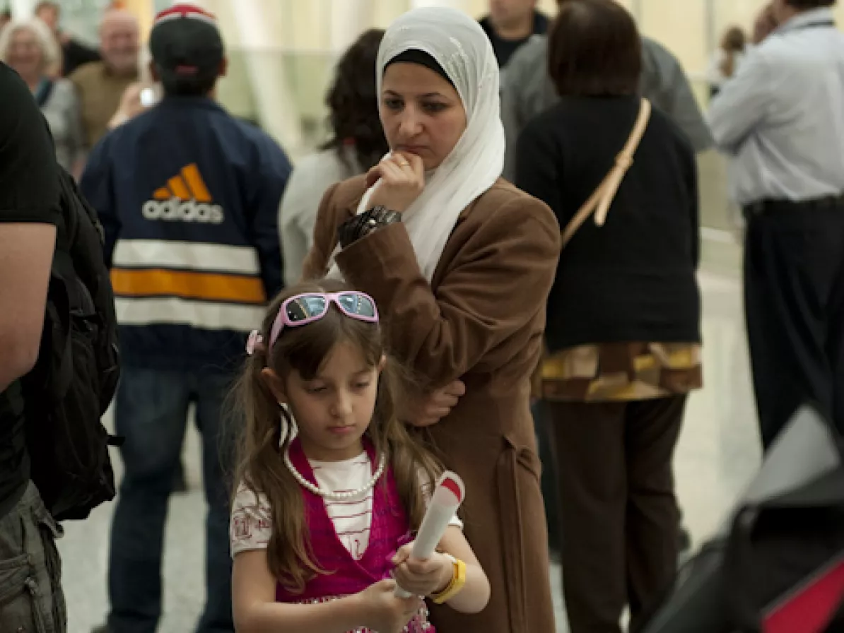 A woman in a hijab looks down at a girl who is rolling up some paper. They are in an airport