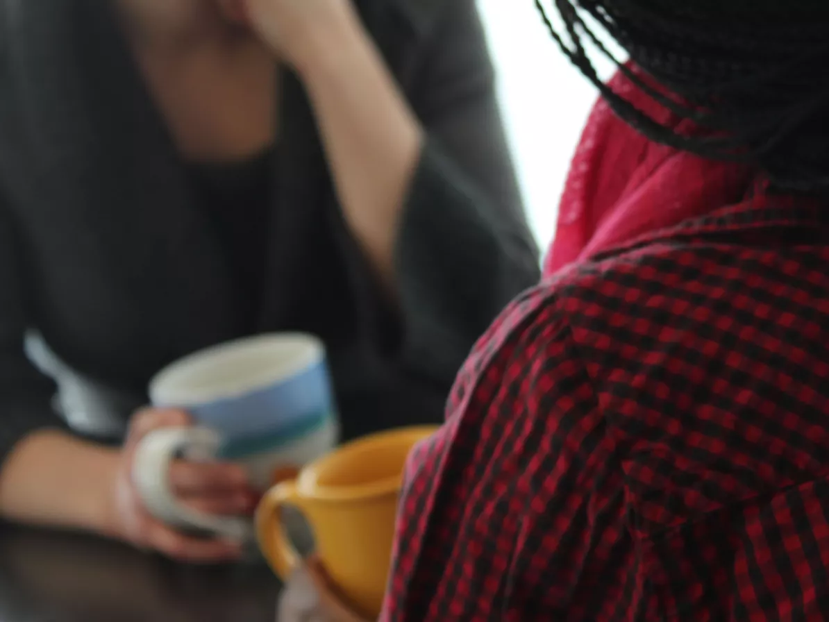 Two people sit at a table facing each other. They are each holding a mug.