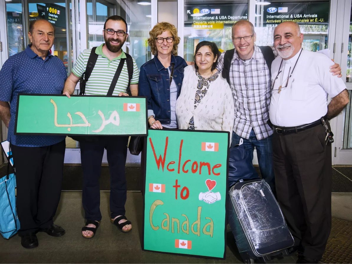 A group of six people with their arms around each other. One is holding a sign that reads, "Welcome to Canada."