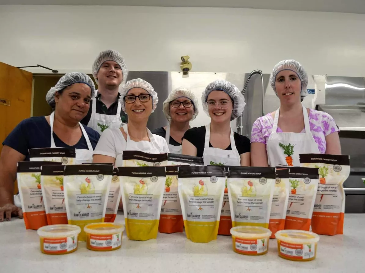 A group of six cooks in hair nets stand behind a table where bags of soup sit.