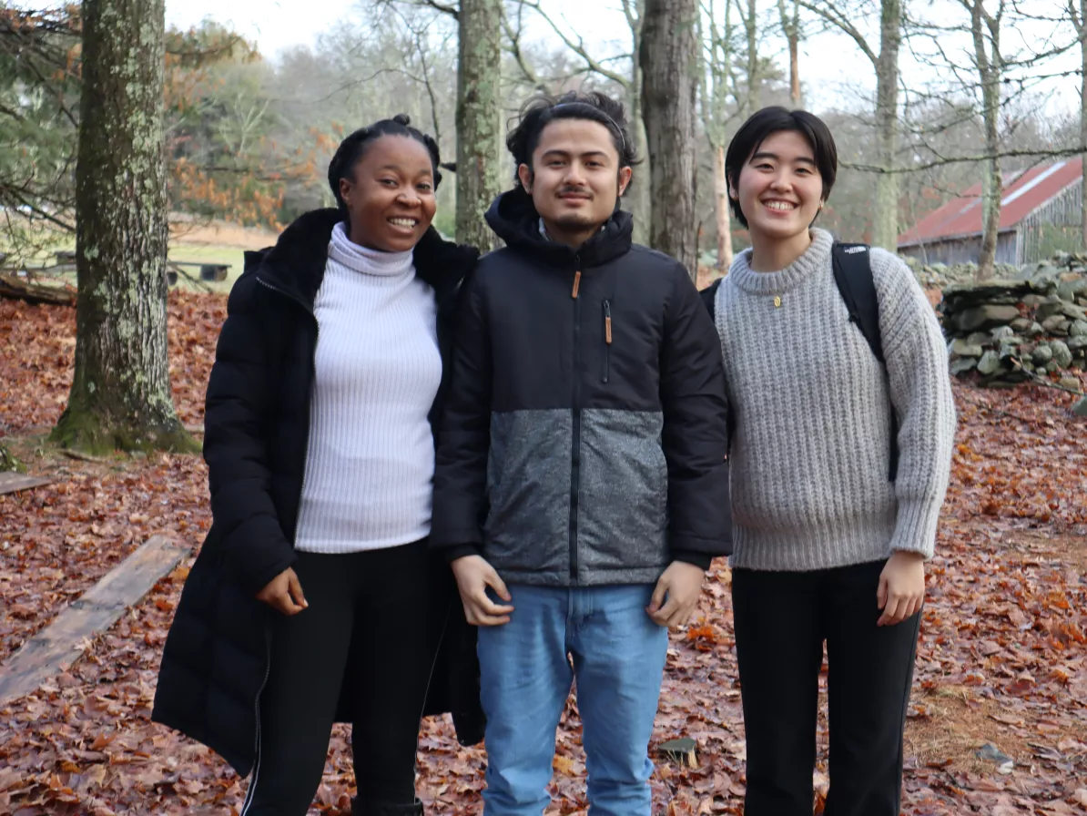 Three young adults stand together and pose for the camera. They are in a wooded area.