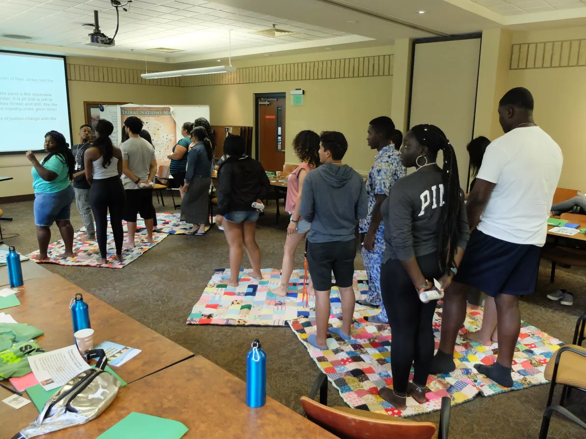 A group of people stand without their shoes on colorful blankets