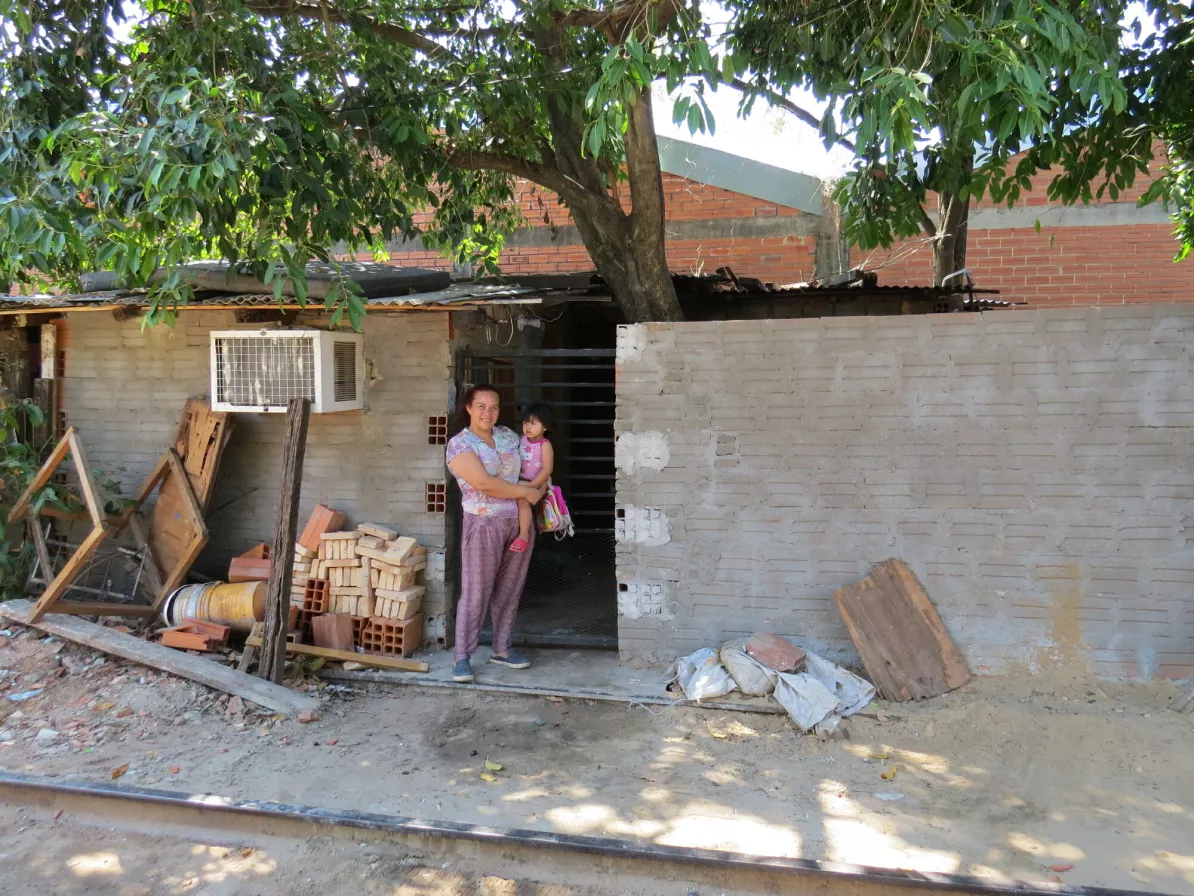A woman holds her child in front of her cement block home