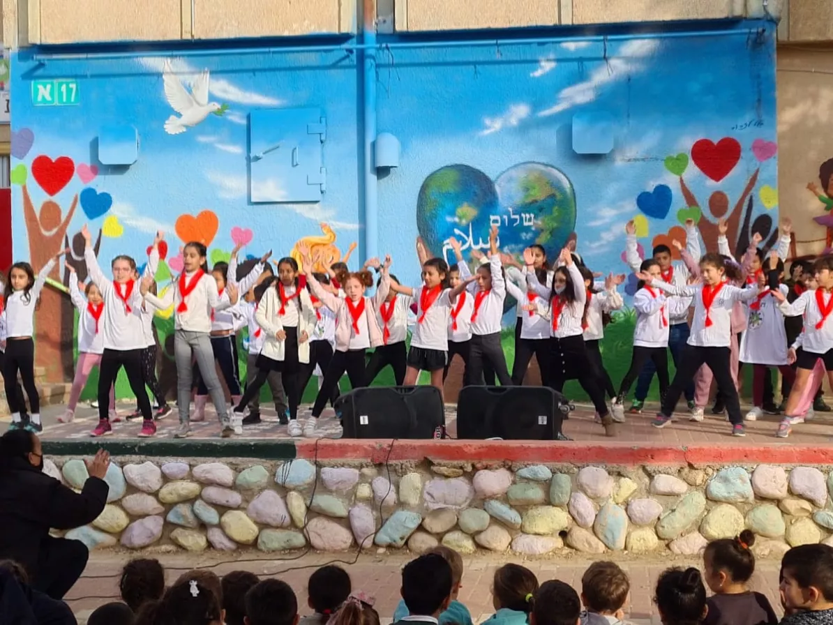 A large group of children wearing red scarves stand on a stage and perform. Many are waving their arms in the air.