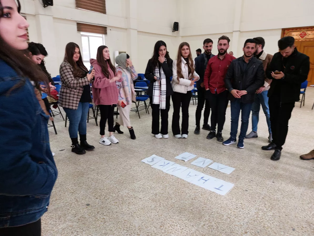 Young adults in Iraq stand in a semi circle around pieces of paper that are on the floor.