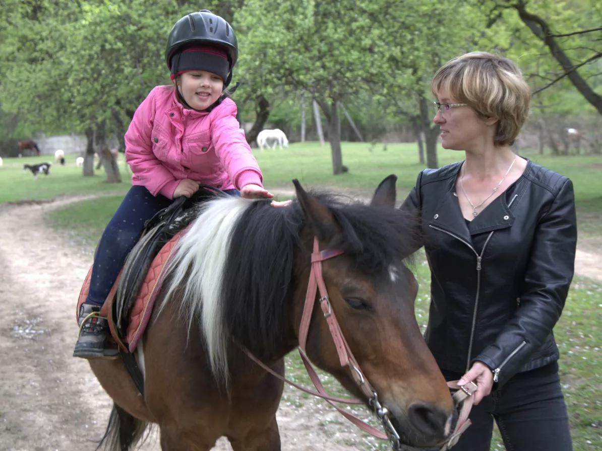 A child in a pink jacket and riding helmet sit a top a horse while a woman guides it.
