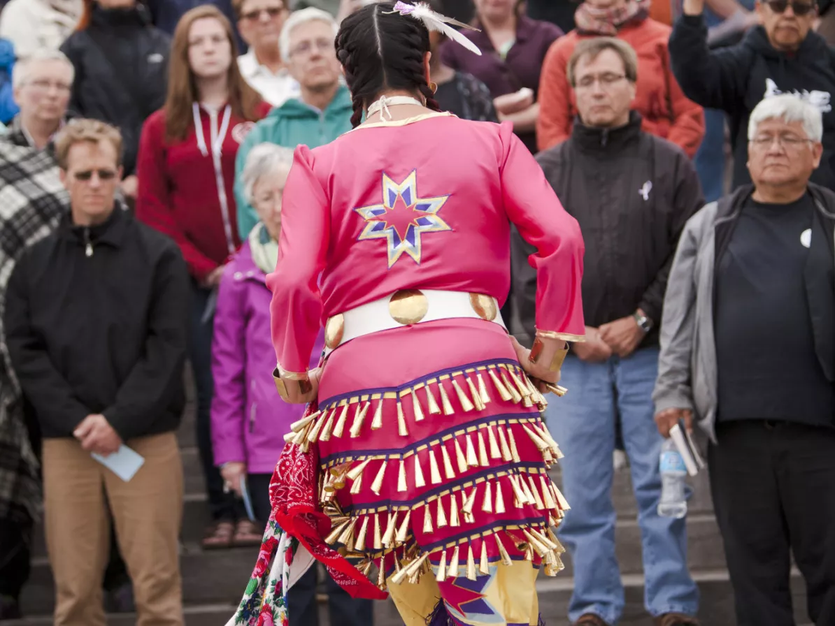 An Indigenous woman in a jingle dress dances in front of a crowd