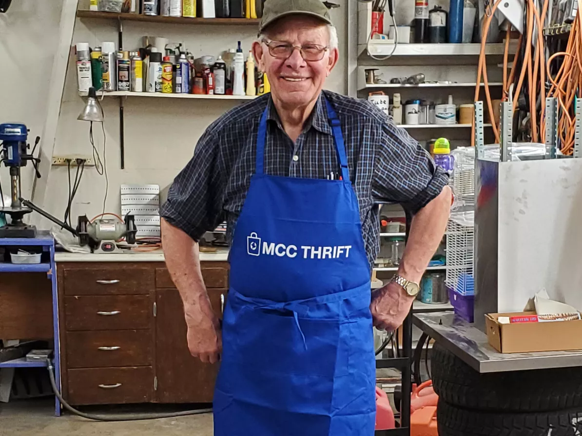 An older man in a ball cap and a blue apron stands in a backroom of a thrift shop