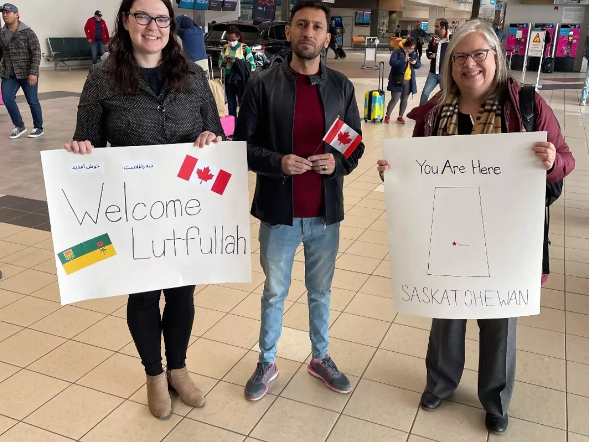 Three people stand together at an airport. Two of them are holding welcome posters
