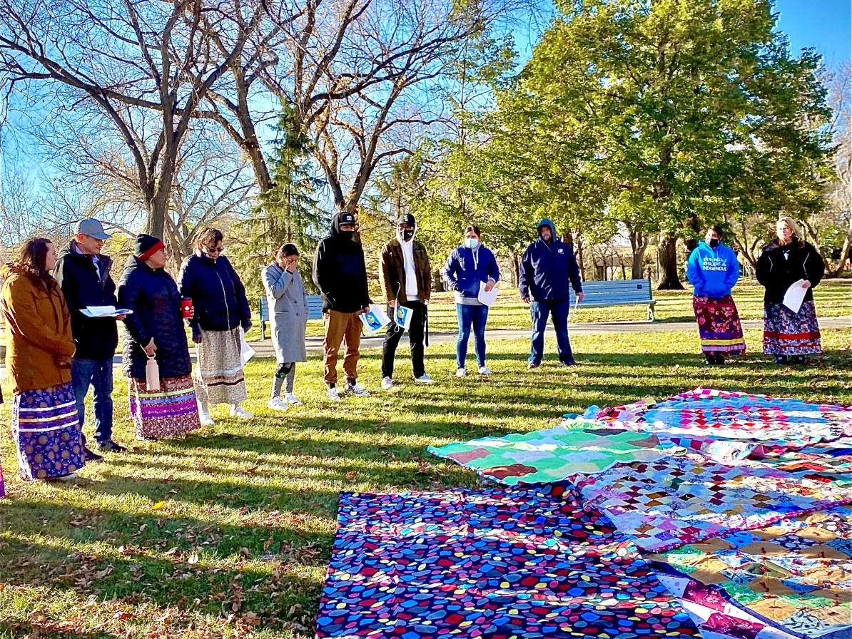 A group of people stand in a circle. In the middle of the circle are quilts spread out on the grass