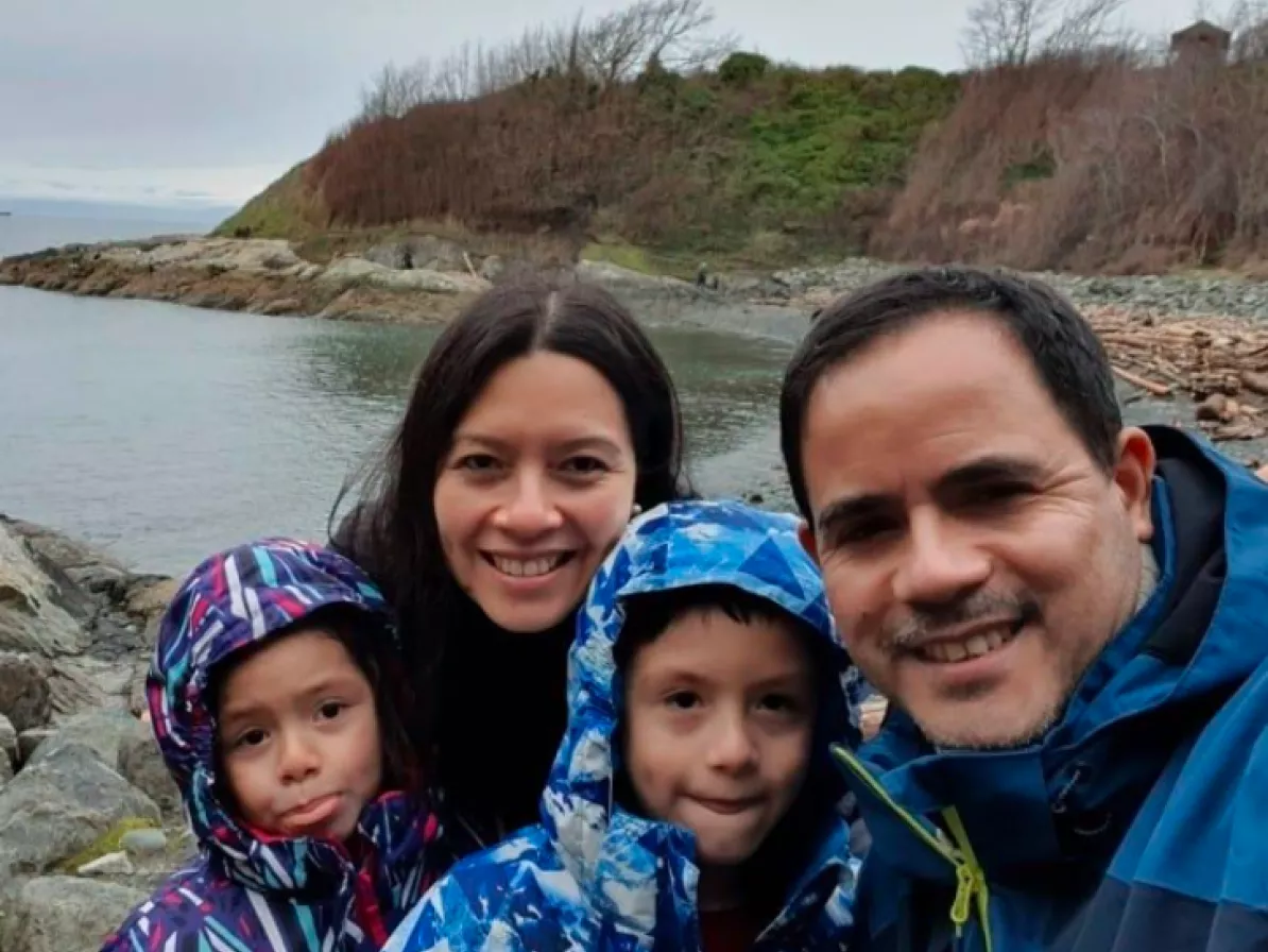 A picture of a family of four: a mother, father, son and daughter in front of a body of water and trees in the backgorund