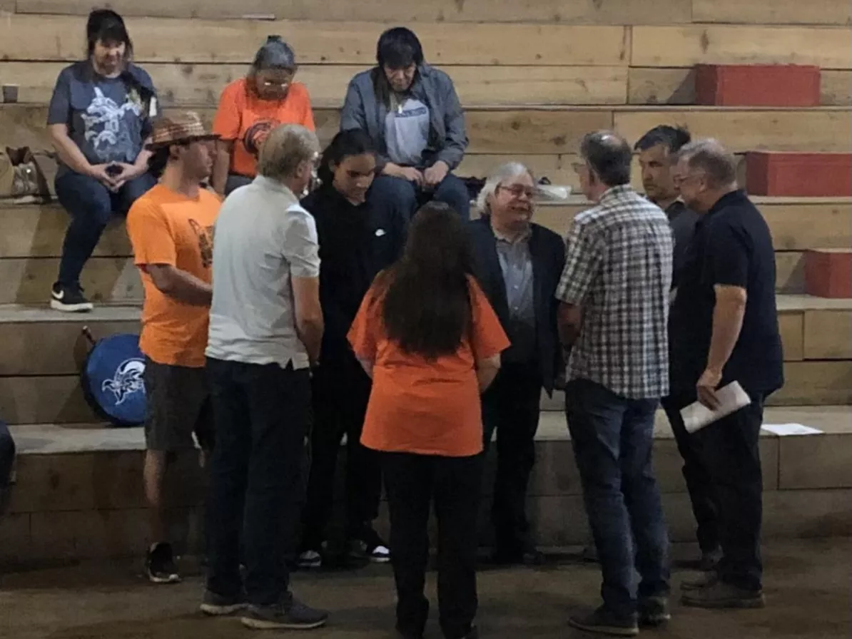 A group of people gathered in a circle praying with a wooden longhouse wall in the background.