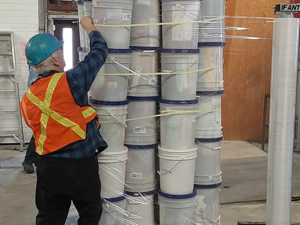 A man wearing a hard hat reaches to fasten together a skid of white buckets.