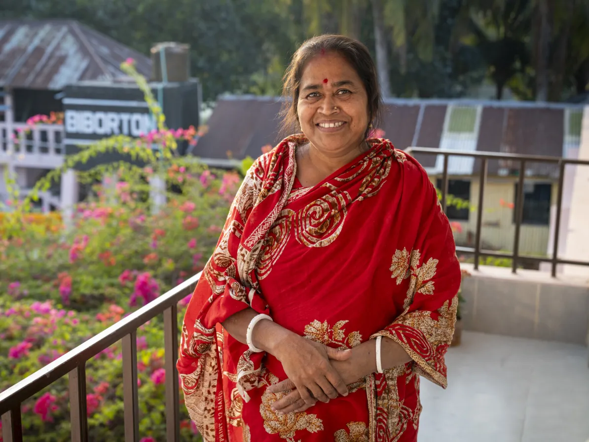 A woman stands on a balcony, smiling. She wears a patterned garment and is surrounded by greenery and flowers in the background.