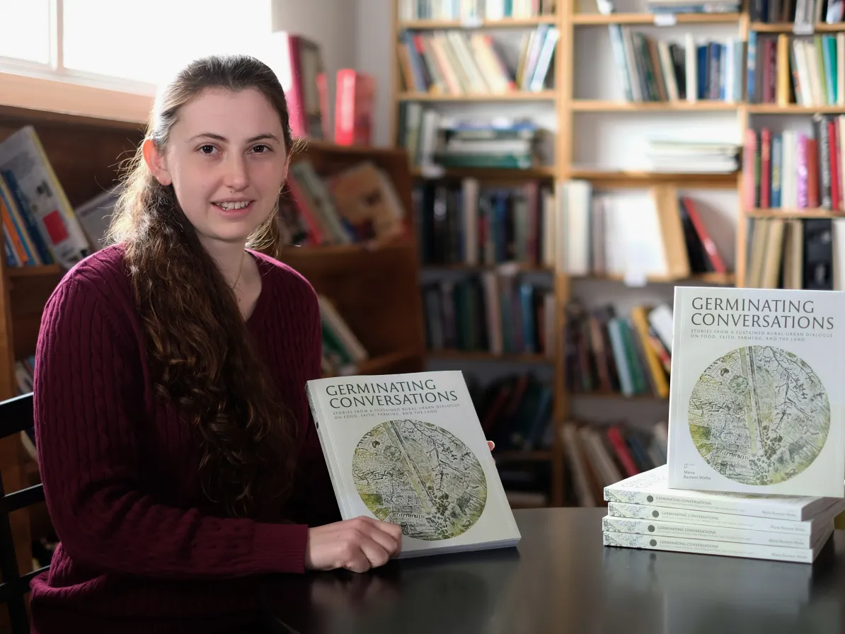 Marta Bennett Wiebe sits at a table holding a copy of the Germinating Conversations book, next to a stack of other copies