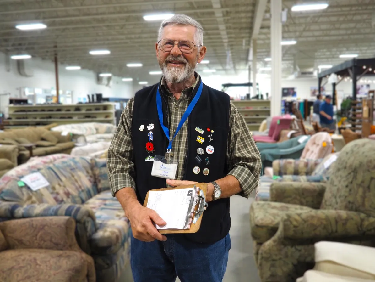 A thrift volunteer, Vern Peters, smiles, holding a clipboard in the furniture department, with couches in the background