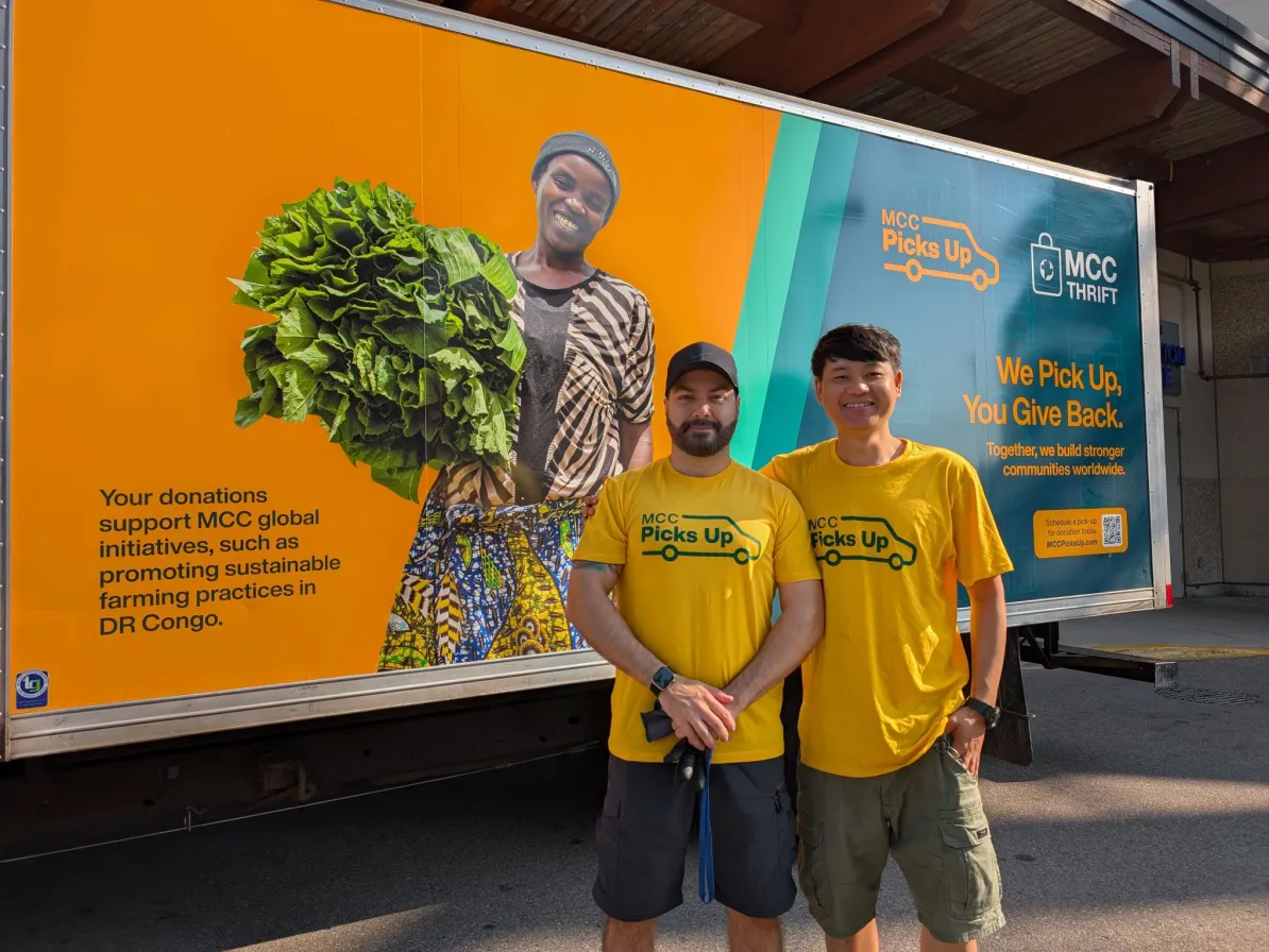 Two men in yellow stand in front of an MCC Thrift truck that is also yellow with images on it.