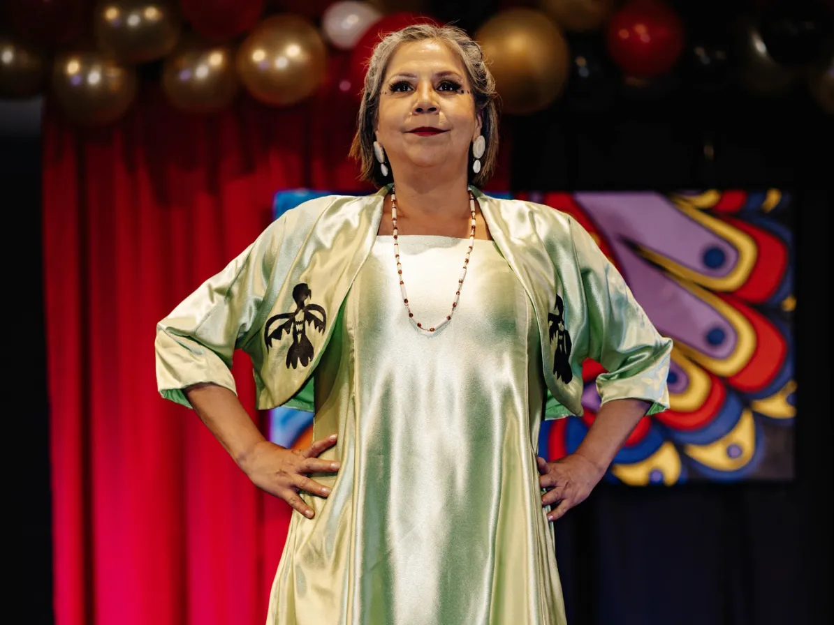 A woman wearing a necklace and earrings stands on a stage of a fashion show. Balloons are in the background.