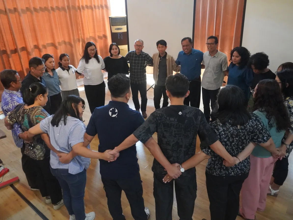A group of people stands in a circle, holding hands, in a spacious room with curtains and a projector screen in the background.